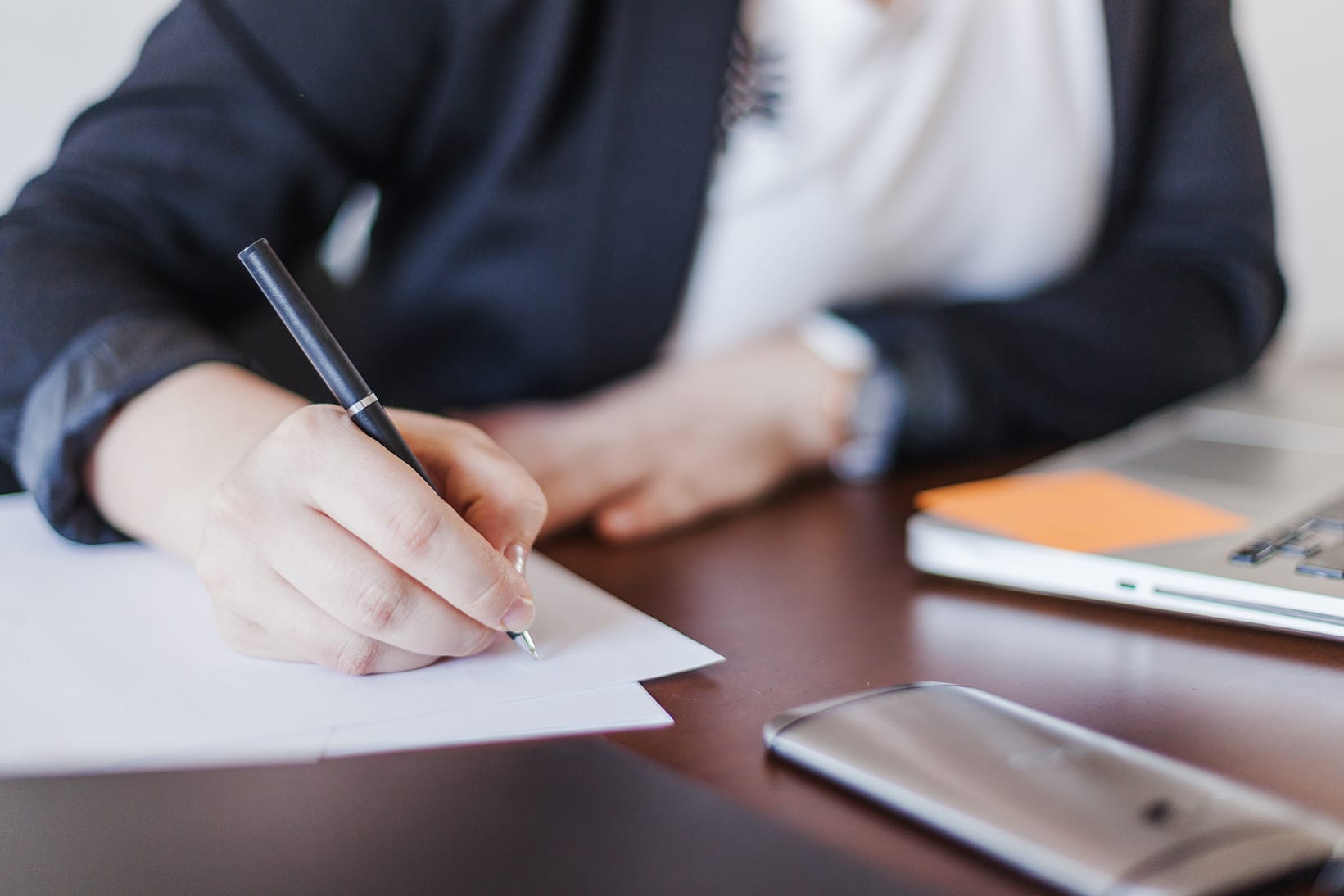 Gros plan sur les mains d'une personne en veste sombre écrivant au stylo sur une feuille blanche. Un ordinateur portable, un carnet orange et un smartphone sont posés sur le bureau en bois sombre à l'arrière-plan, légèrement flous.