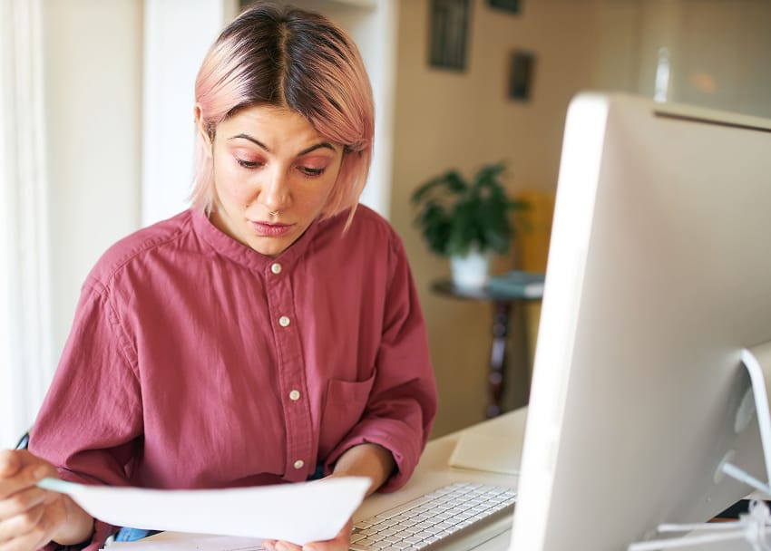 Femme aux cheveux roses concentrée, consultant un document papier devant un écran d'ordinateur dans un espace de travail à domicile lumineux — illustration de la gestion administrative et comptable d'une petite entreprise, d'une entrepreneuse suisse